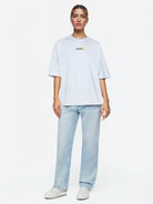 Eine Person steht mit dem Blick nach vorne und trägt das trueprodigy Juniper F Oversize-Damen-T-Shirt mit Logo-Smile-Grafik in Hellgrau, gepaart mit hellblauen Jeans und weißen Turnschuhen, vor einem schlichten weißen Hintergrund.