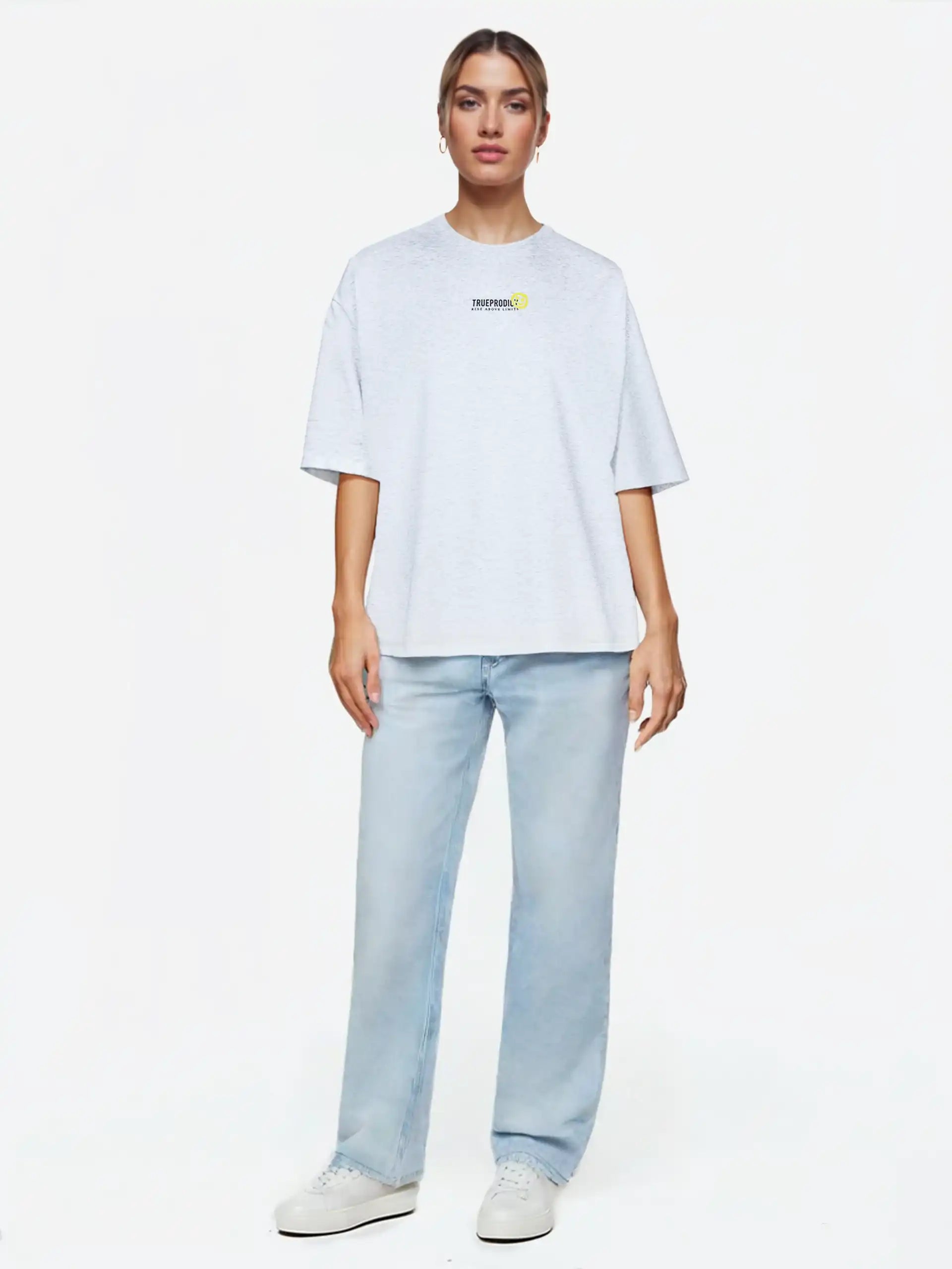 Eine Person steht mit dem Blick nach vorne und trägt das trueprodigy Juniper F Oversize-Damen-T-Shirt mit Logo-Smile-Grafik in Hellgrau, gepaart mit hellblauen Jeans und weißen Turnschuhen, vor einem schlichten weißen Hintergrund.