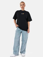 Vor einem schlichten weißen Hintergrund trägt eine Person das trueprodigy Juniper F Oversize-Damen-T-Shirt mit Logo-Smile-Grafik auf der Vorderseite in Schwarz, gepaart mit hellblauen Röhrenjeans und weißen Sneakers.