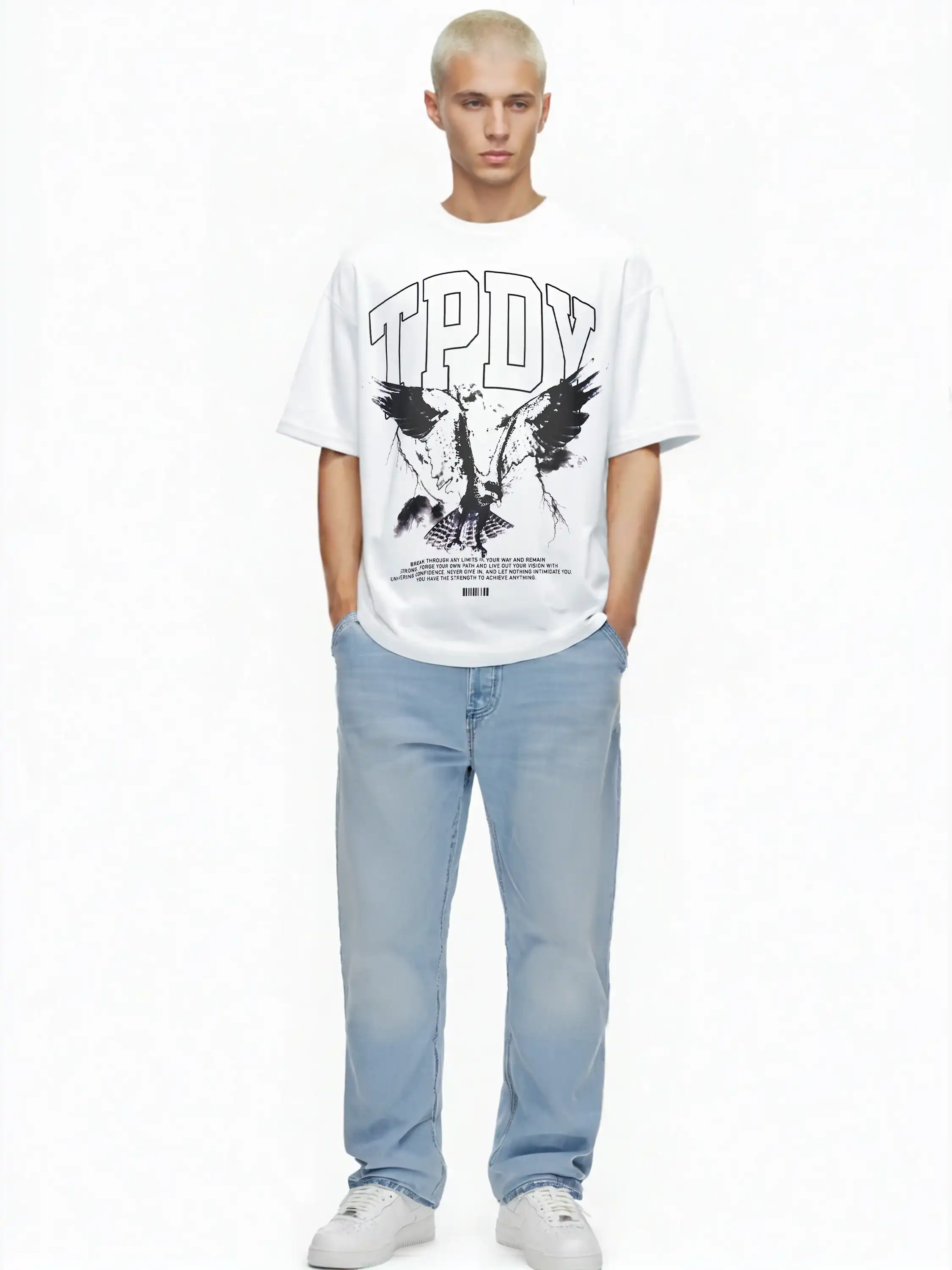 Eine Person mit kurzen blonden Haaren trägt das trueprodigy Yuki Oversize-T-Shirt mit Adlergrafik & TPDY-Schriftzug, hellblaue Jeans und weiße Turnschuhe und steht mit den Händen in den Taschen vor einem schlichten weißen Hintergrund.