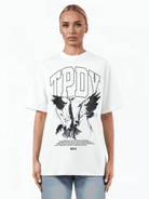 Eine Person steht vor einem schlichten weißen Hintergrund und trägt das TrueProdigy Yuki F Unisex T-Shirt mit dem Schriftzug „TPDY“ oben und einem kunstvollen Adlermotiv darunter. Der übergroße Schnitt passt perfekt zu den zurückgekämmten Haaren und der hellblauen Jeans.