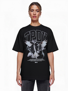 Eine Person steht mit Blick nach vorne in einem trueprodigy Oversize-T-Shirt mit Adlergrafik & TRUEPRODIGY-Schriftzug: loser schwarzer Jersey, "TPDY" in großen weißen Buchstaben, Vogelgrafik mit ausgebreiteten Flügeln, kleiner weißer Text und ein Barcode am unteren Rand.