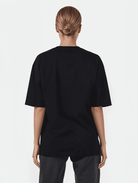 Person mit zurückgebundenem Haar, die ein übergroßes Sam F-T-Shirt von Trueprodigy und dunkle Hosen trägt und vor einem weißen Hintergrund mit dem Gesicht nach hinten blickt.