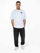 Ein Mann steht vor einem schlichten weißen Hintergrund und trägt das trueprodigy Oversize-T-Shirt mit Spray-Smiley & Rückenprint "Happy Mind" mit kleiner blauer Brustgrafik, gepaart mit schwarzer Jeans und weißen Turnschuhen, die Hände in den Taschen, den Blick nach vorne gerichtet.