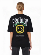 Eine Person mit hellbraunen, nach hinten gebundenen Haaren trägt das trueprodigy Oversize-T-Shirt mit Spray-Smiley & Happy-Mind-Print. Das schwarze Unisex-Shirt zeigt "TRUE PRODIGY" in weiß, einen gelben Spray-Smiley und einen grünen "HAPPY MIND"-Text auf dem Rücken.