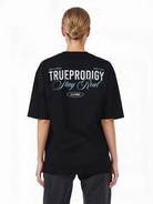 Eine Person mit blonden Haaren in einem tiefen Dutt ist von hinten zu sehen. Sie trägt das trueprodigy Oversize-T-Shirt mit "Stay Real" Schriftzug vorne & hinten, mit weißem und hellblauem Text auf schwerem Jerseystoff.