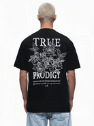 Eine Person mit kurzen Haaren blickt abgewandt und trägt ein schwarzes Unisex-T-Shirt mit dem Namen Alex von Trueprodigy. Es zeigt weiße Blumenmuster und „True Prodigy“ auf dem Rücken, darunter ein motivierender Spruch. Es passt perfekt zu hellblauen Jeans.