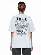 Eine Person mit braunem Haar trägt ein übergroßes hellgraues Unisex-T-Shirt „Alex F“ von trueprodigy mit dem Schriftzug „TRUE PRODIGY“ und einer Rosengrafik auf der Rückseite. Darunter befindet sich ein kleinerer Text. Der Look wird durch eine dunkle Hose aus bequemem Jerseystoff abgerundet.