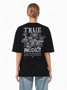 Mit hellbraunem Haar blickt eine Person in einem übergroßen schwarzen Alex F T-Shirt von trueprodigy abgewandt in die Kamera. Es besteht aus Jerseystoff und zeigt weiße Blumenmuster und einen inspirierenden Text. Kombiniert mit hellblauen Jeans strahlt es lässiges Selbstbewusstsein aus.