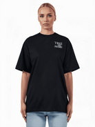 Vor einem schlichten Hintergrund steht eine Person, die ein übergroßes Alex F-T-Shirt mit dem „Trueprodigy“-Logoaufdruck auf der Vorderseite und hellblaue Jeans trägt.