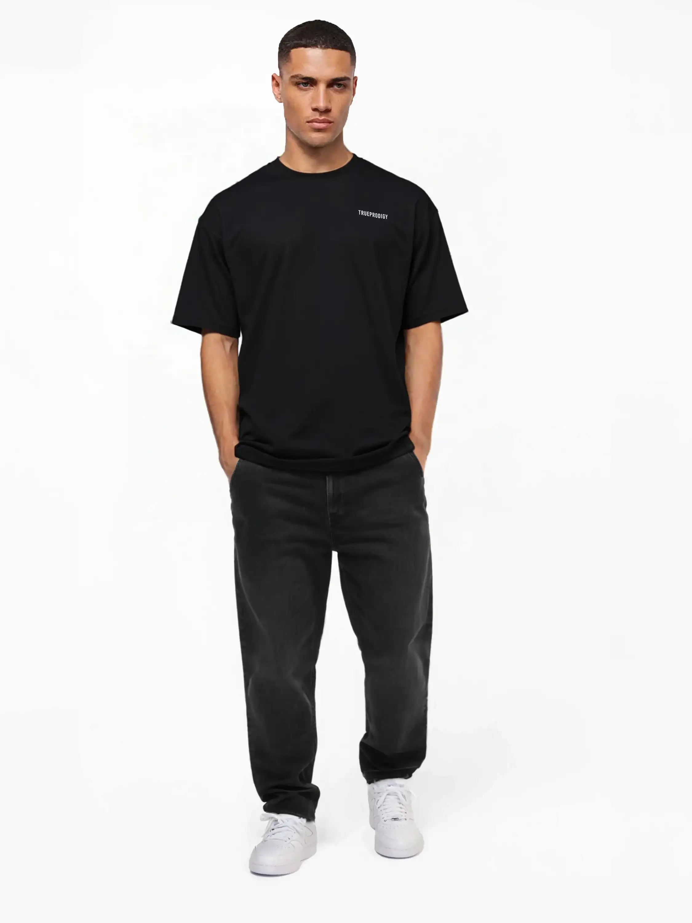 Eine Person steht mit dem Gesicht nach vorne vor einem schlichten weißen Hintergrund und trägt das trueprodigy Lior Oversize-T-Shirt mit Brustlogo und Rückenprint "never back down", dunkle Jeans und weiße Turnschuhe mit den Händen in den Taschen.