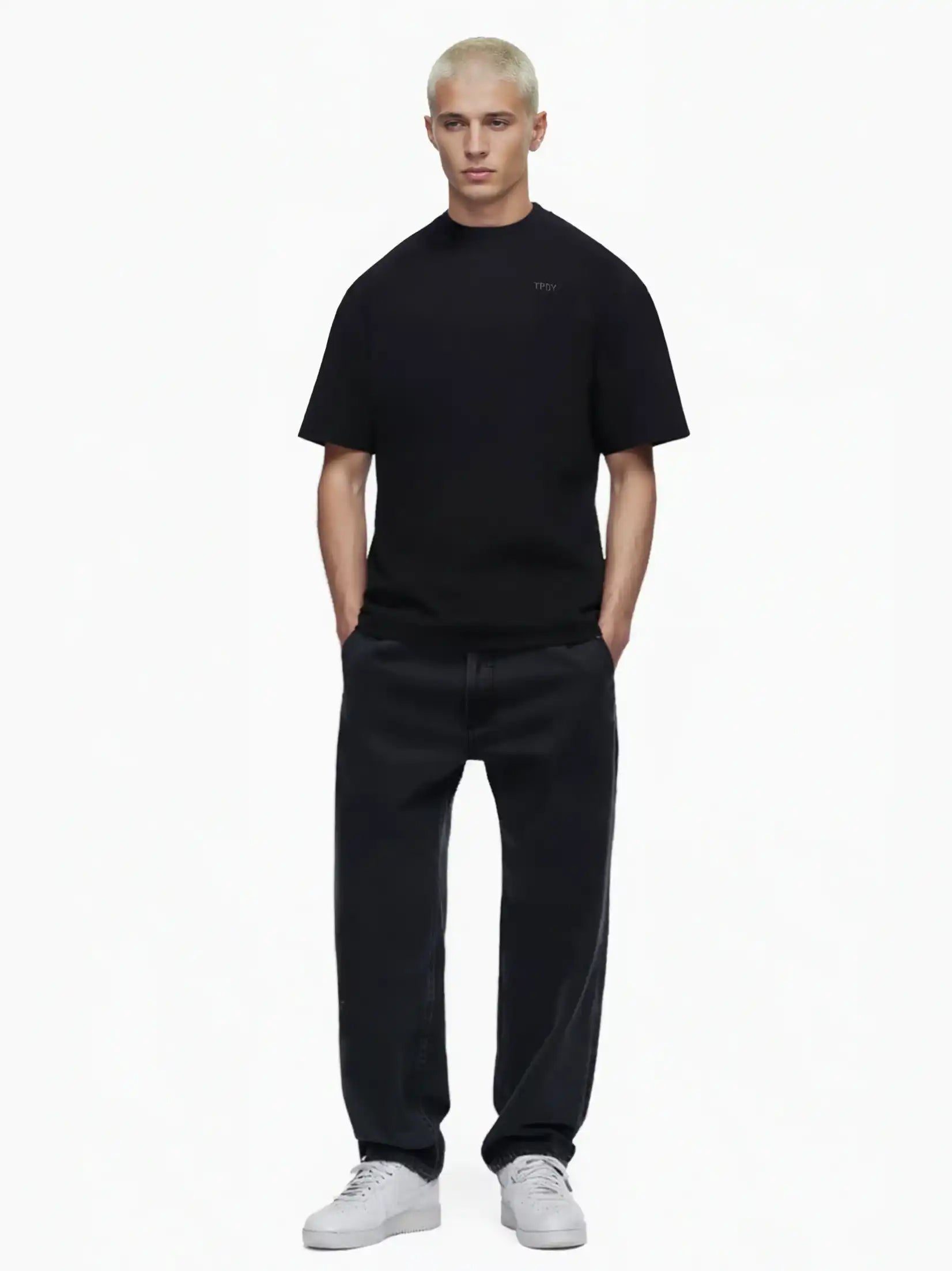 Eine Person mit kurzen, gebleichten blonden Haaren trägt das Trueprodigy Haram Oversize-Herren-T-Shirt mit dezentem TPDY-Sticklogo in Schwarz, weite schwarze Jeans und weiße Turnschuhe. Sie steht mit den Händen in den Taschen vor einem schlichten weißen Hintergrund.