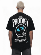 Eine Person mit kurzen Haaren blickt abgewandt und trägt das Kenji Oversized T-Shirt von trueprodigy. Es ist schwarz und trägt die Aufschrift „TRUE PRODIGY“ und „KEEP SMILING“ sowie die Grafik eines lächelnden Gesichts mit X-Augen und blauen Tropfen – ein trendiger Unisex-Stil.