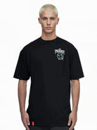 Eine Person mit kurzen blonden Haaren trägt ein Kenji Oversized T-Shirt mit Druck von trueprodigy, mit dem „The Prodigy“-Logo und einer kleinen Grafik oben links. Der schwere Jersey bietet eine bequeme Unisex-Passform vor dem schlichten weißen Hintergrund.