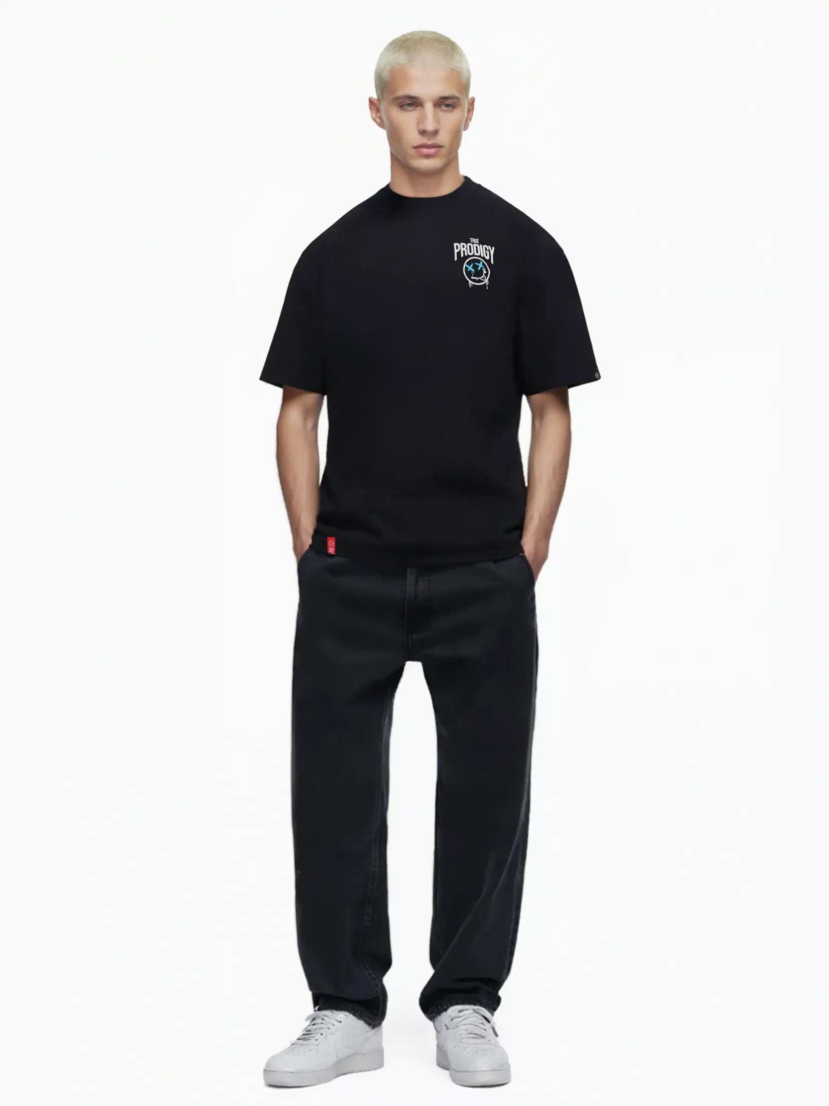 Eine Person mit kurzen blonden Haaren trägt ein Kenji Oversized T-Shirt mit Druck von trueprodigy, kombiniert mit einer dunklen Hose vor einem schlichten weißen Hintergrund. Knackig weiße Sneakers runden den Look ab.