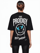 Eine Person mit braunen Haaren blickt abgewandt und trägt das Kenji Oversized T-Shirt von trueprodigy. Es trägt den Schriftzug „TRUE PRODIGY“ in Weiß, ein stilisiertes Gesicht mit blauem Kreuz und dem Schriftzug „KEEP SMILING“. Dieses Unisex-Shirt passt perfekt zu der stylischen schwarzen Hose.