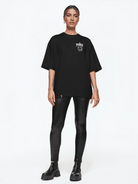Eine Person steht vor einem weißen Hintergrund und trägt das Kenji Oversized T-Shirt mit Druck von trueprodigy, dazu schwarze Leggings und klobige Stiefel. Die zurückgekämmten Haare unterstreichen den neutralen Ausdruck und vereinen mühelos Stil und Komfort.