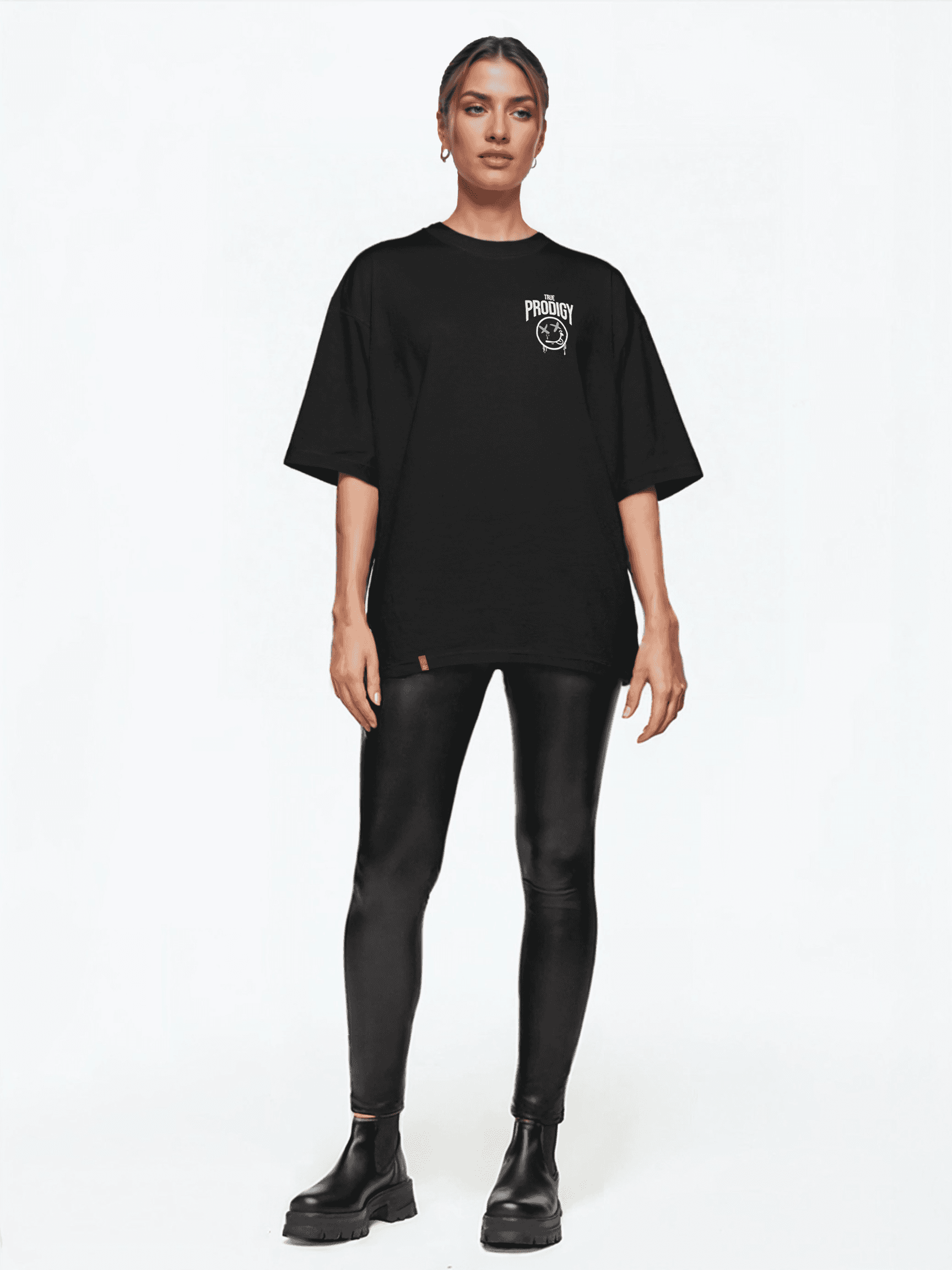 Eine Person steht vor einem weißen Hintergrund und trägt das Kenji Oversized T-Shirt mit Druck von trueprodigy, dazu schwarze Leggings und klobige Stiefel. Die zurückgekämmten Haare unterstreichen den neutralen Ausdruck und vereinen mühelos Stil und Komfort.