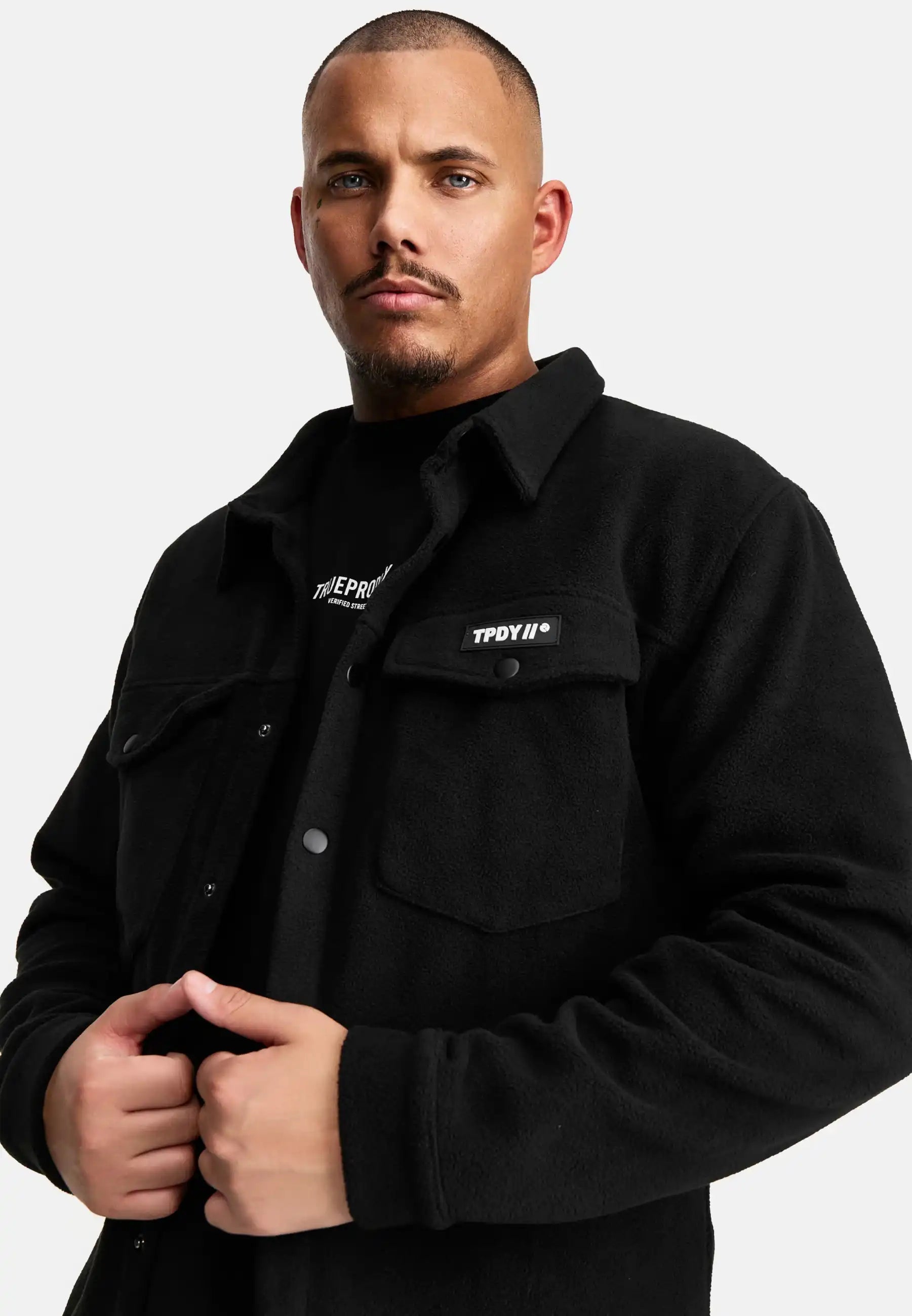 Ein Mann mit kurzgeschnittenem Haar und gestutztem Bart trägt das schwarze trueprodigy Bill Fleece Oversized Hemd mit Druckknöpfen und einer Brusttasche mit TPDYIN-Aufnäher über einem schwarzen Hemd auf weißem Grund.