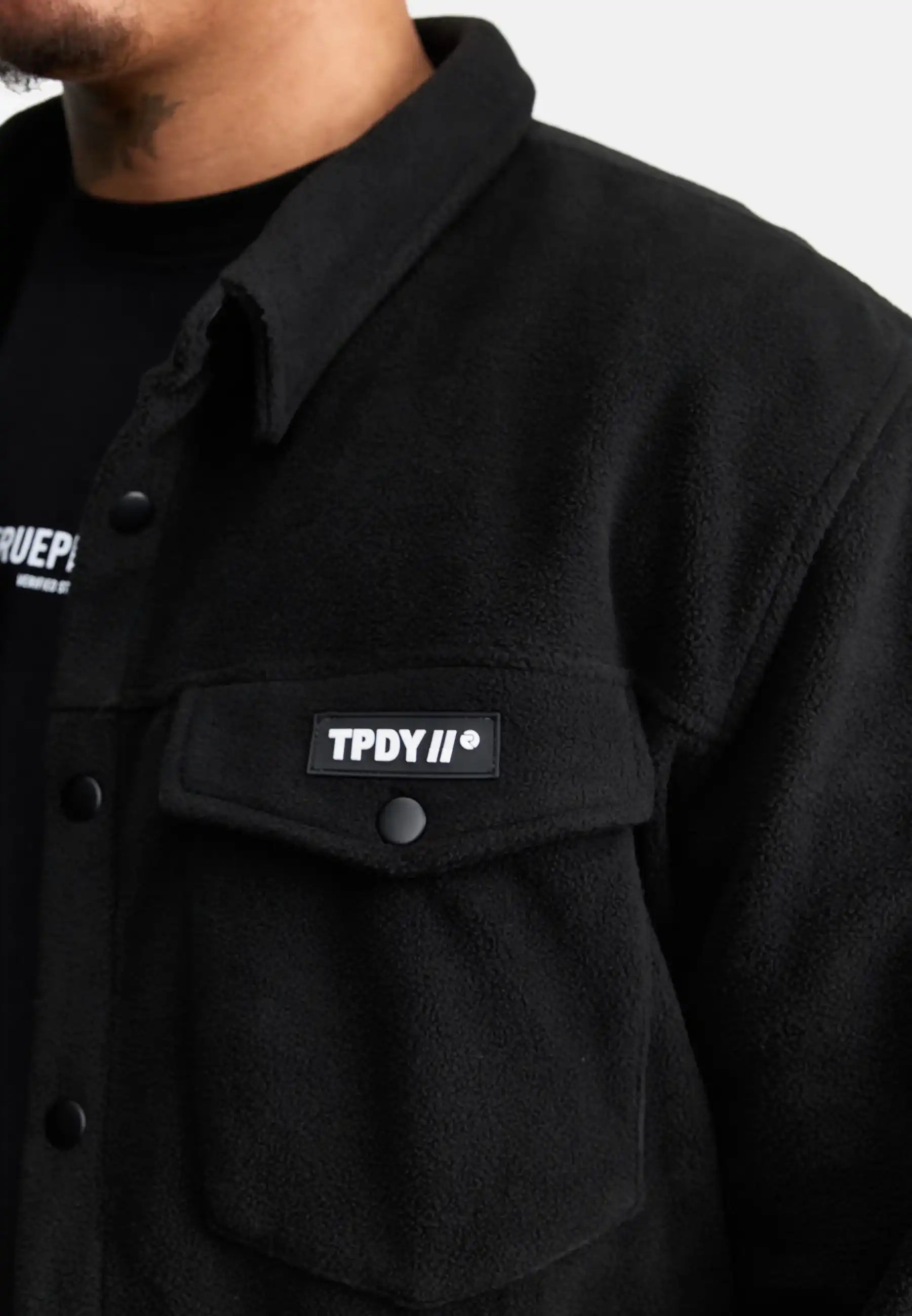 Eine Person trägt das trueprodigy Bill Fleece Oversized Hemd in Schwarz mit einer geknöpften Brusttasche mit einem rechteckigen TPDY II®-Aufnäher, der über einen weiteren übergroßen schwarzen Saum mit weißem Text gelegt ist.