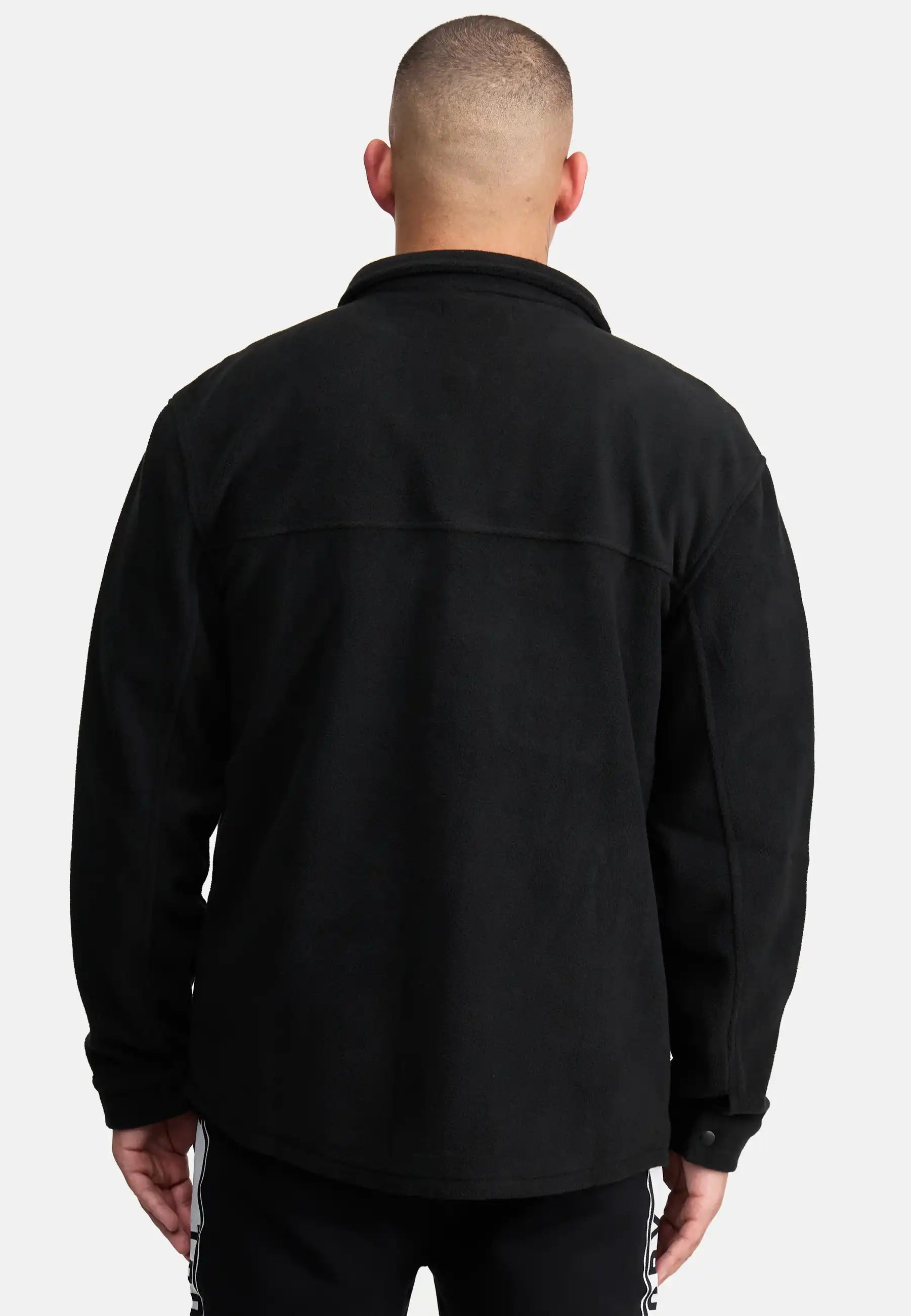 Ein Mann mit kurzen Haaren ist von hinten zu sehen und trägt ein schwarzes Trueprodigy Bill Fleece Oversized Hemd und eine schwarze Hose mit weißen Seitenstreifen. Er steht vor einem schlichten hellgrauen Hintergrund, der einen stilvollen, unaufdringlichen Look erzeugt.