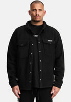 Ein Mann blickt nach vorne in einem schwarzen Trueprodigy Bill Fleece Oversized Hemd über einem schwarzen T-Shirt. Das Hemd hat zwei Brusttaschen und ein kleines rechteckiges Etikett auf der linken Seite, vor einem schlichten weißen Hintergrund.