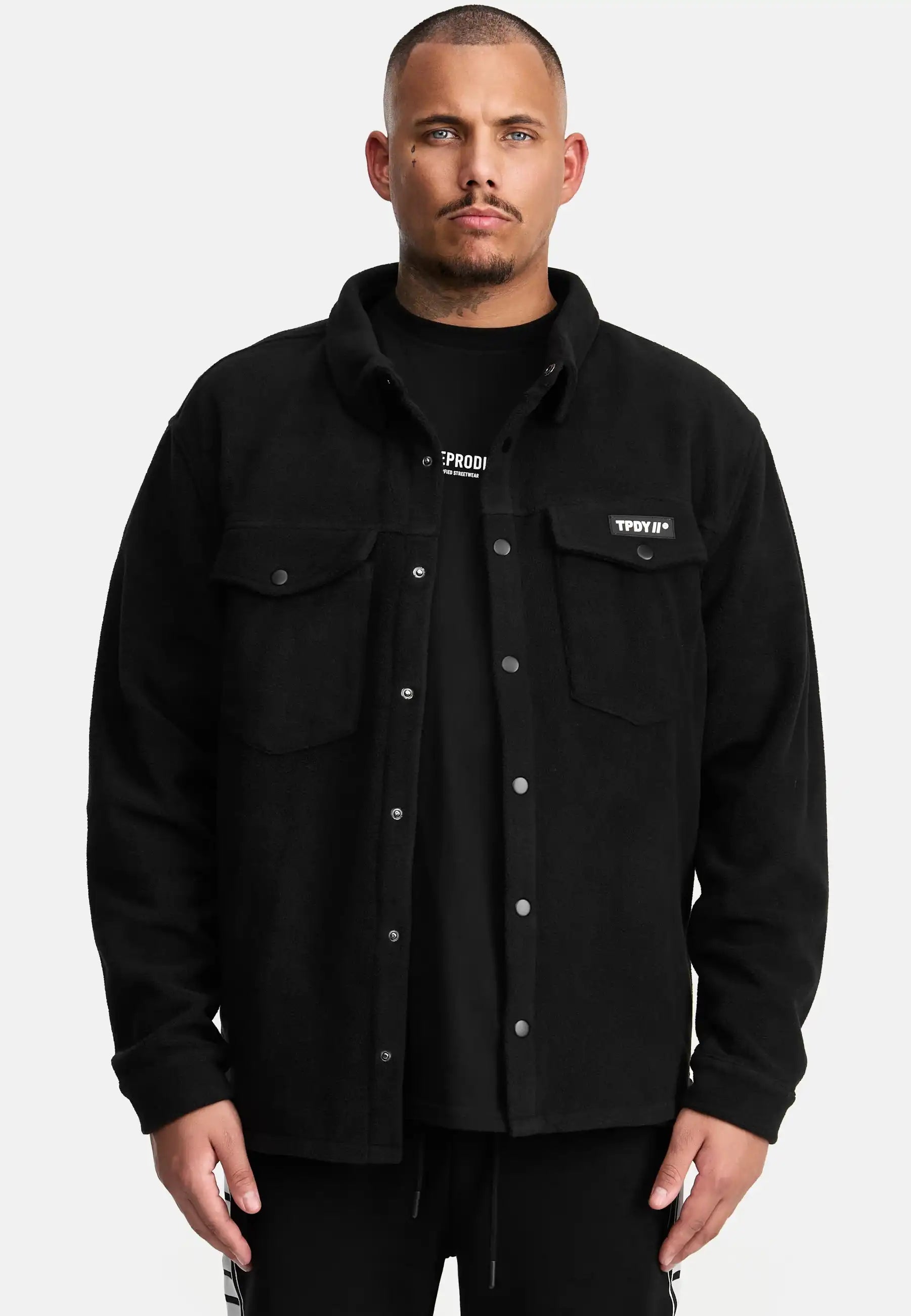 Ein Mann blickt nach vorne in einem schwarzen Trueprodigy Bill Fleece Oversized Hemd über einem schwarzen T-Shirt. Das Hemd hat zwei Brusttaschen und ein kleines rechteckiges Etikett auf der linken Seite, vor einem schlichten weißen Hintergrund.