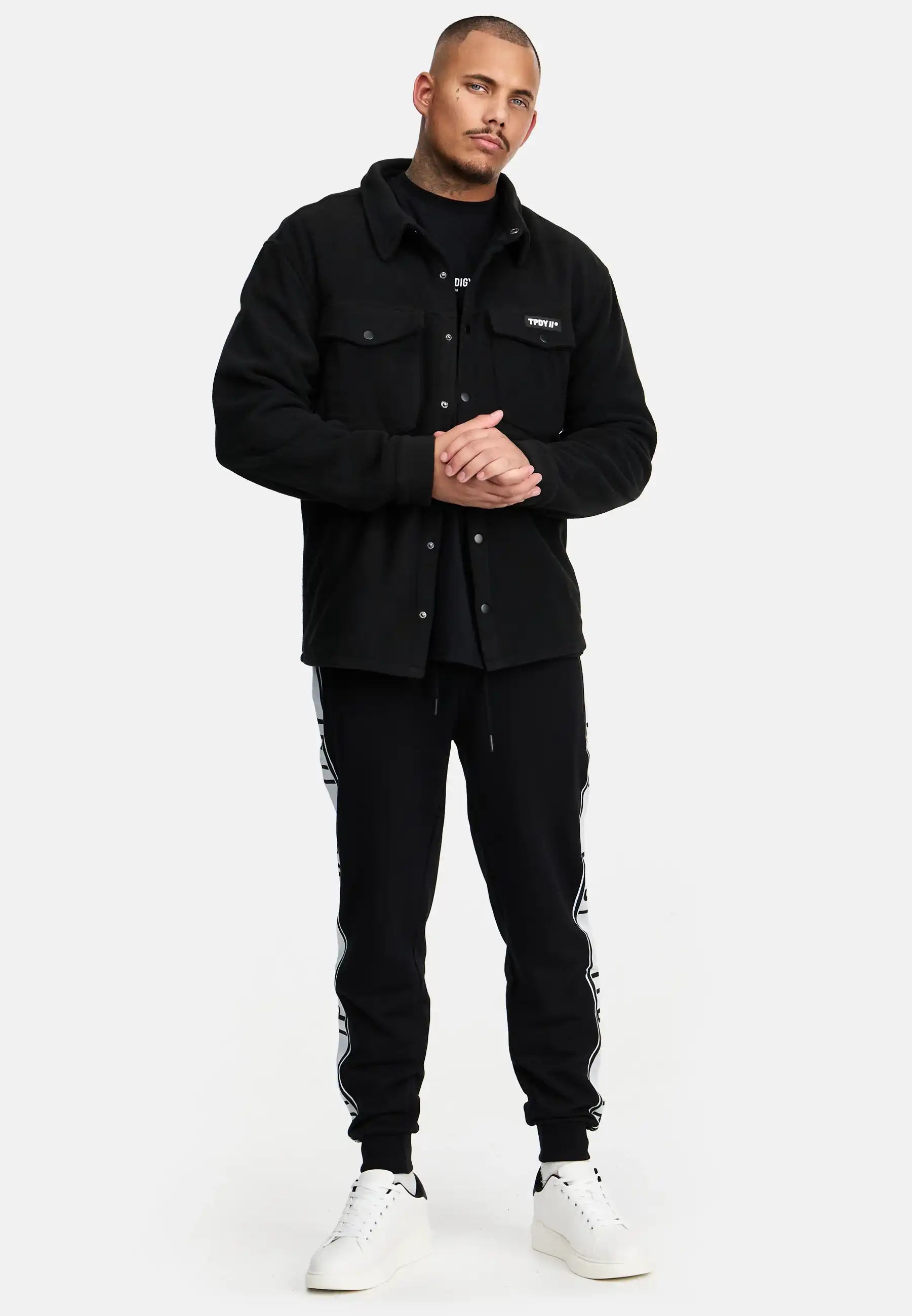 Ein Mann steht vor einem schlichten weißen Hintergrund und trägt das trueprodigy Bill Fleece Oversized Hemd in Schwarz über einem schwarzen Hemd, gepaart mit einer schwarzen Jogginghose mit weißen Seitenstreifen und weißen Sneakers. Er blickt nach vorne, die Hände leicht verschränkt.