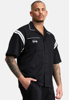 Ein Mann mit kurzen Haaren und Tattoos trägt das trueprodigy Steve Oversized Retro Hemd, ein schwarzes, kurzärmeliges Button-up-Hemd mit weißen Kontraststreifen und Tpdsy-Stickerei auf der Brusttasche, und steht mit einer Hand in der Tasche.
