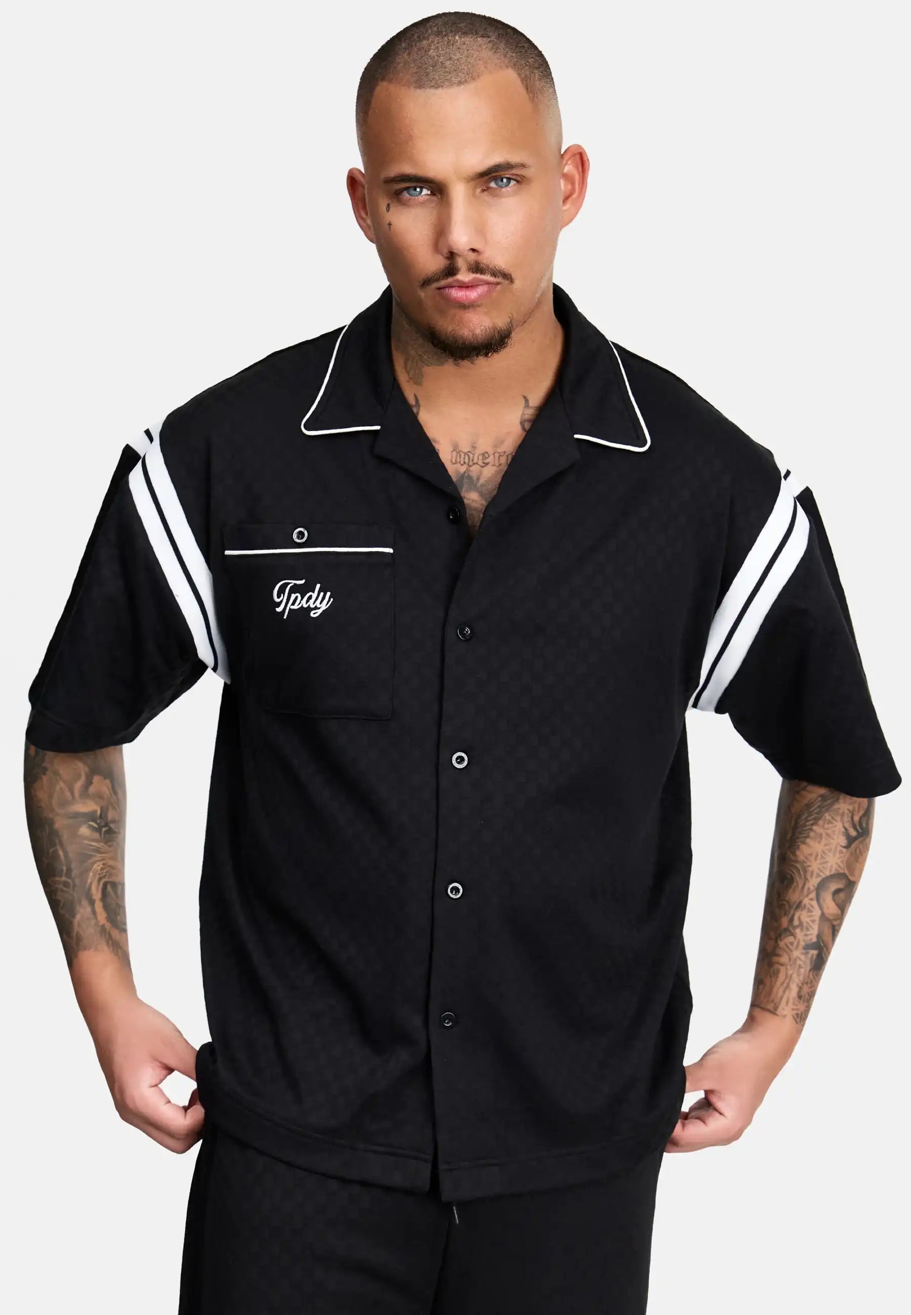 Ein tätowierter Mann mit Kurzhaarschnitt trägt das trueprodigy Steve Oversized Retro Hemd, ein schwarzes, kurzärmeliges Button-up-Hemd mit weißen Kontraststreifen an den Ärmeln, weißer Paspelierung am Kragen und gesticktem "Tpdv" auf der Brusttasche.
