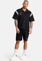 Ein Mann mit Tattoos und Kurzhaarschnitt steht vor einem schlichten Hintergrund und trägt das Steve Oversized Retro Hemd von trueprodigy mit weißen Kontraststreifen an den Schultern, schwarze Shorts, weiße Socken und Turnschuhe.