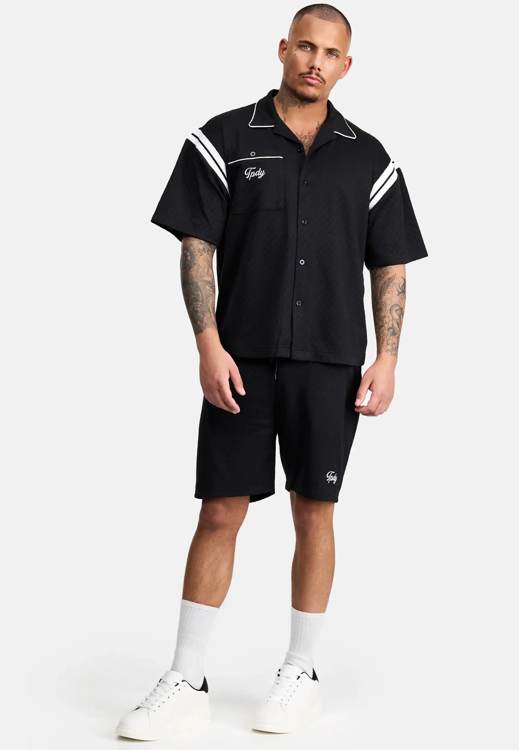 Ein Mann mit Tattoos und Kurzhaarschnitt steht vor einem schlichten Hintergrund und trägt das Steve Oversized Retro Hemd von trueprodigy mit weißen Kontraststreifen an den Schultern, schwarze Shorts, weiße Socken und Turnschuhe.
