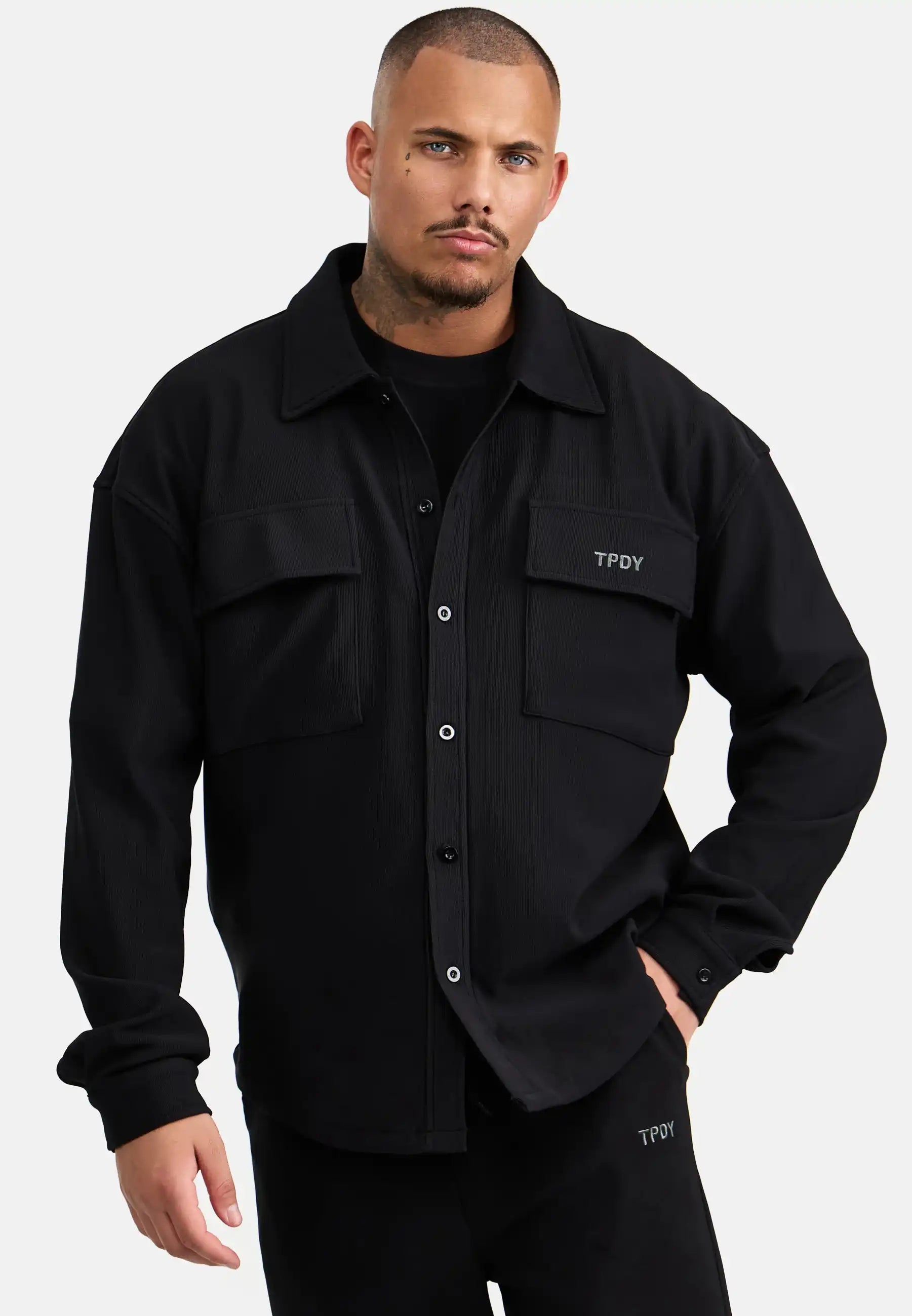 Ein Mann mit kurzen Haaren und gestutztem Bart trägt das trueprodigy Wyatt Oversized Langarm Hemd in Schwarz mit gerippter Struktur und zwei Brusttaschen, gepaart mit einer passenden schwarzen Hose, vor einem schlichten hellen Hintergrund.