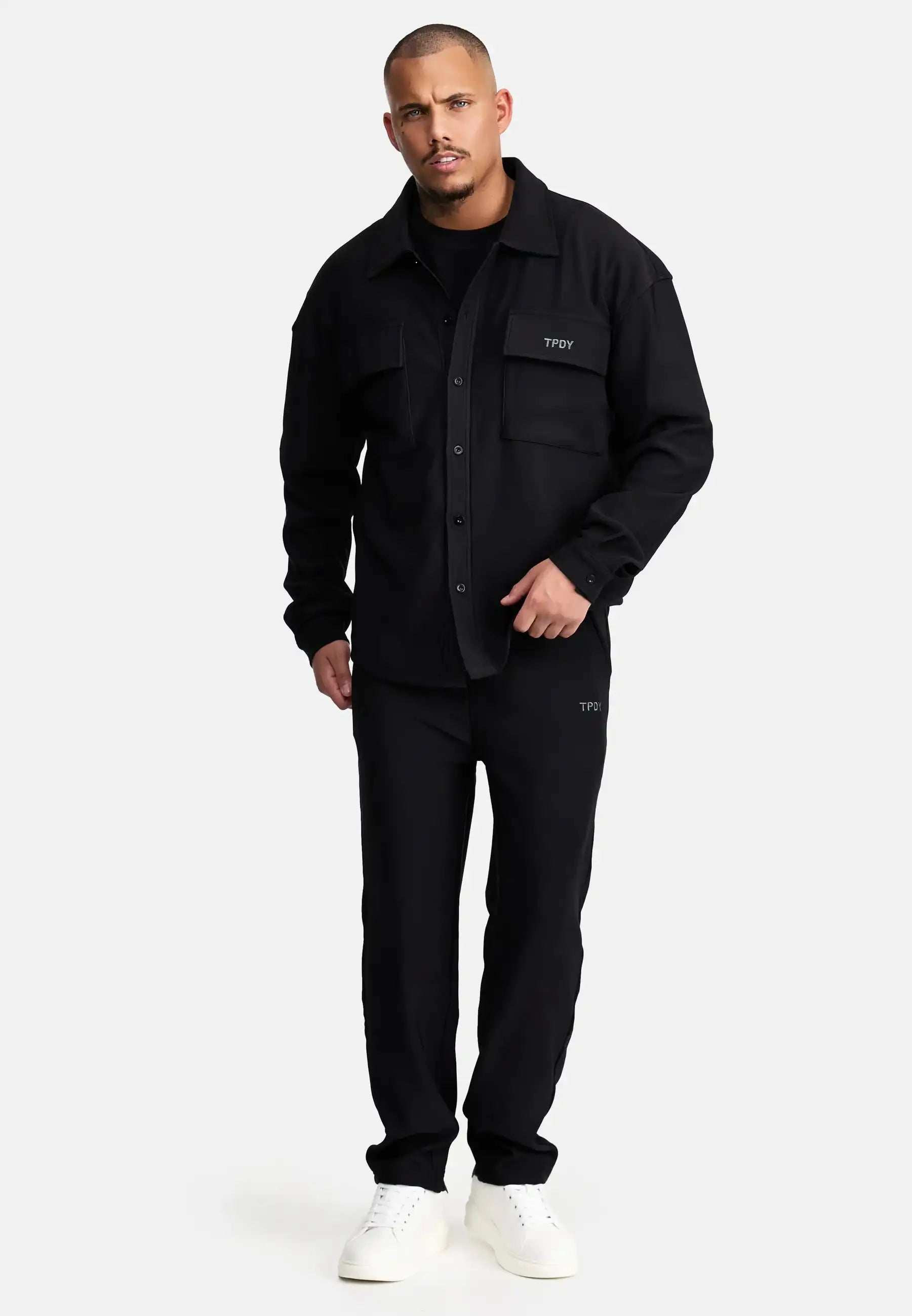 Vor einem schlichten Hintergrund trägt ein Mann mit kurzen Haaren und gepflegtem Bart das Wyatt Oversized Langarm Hemd von trueprodigy, schwarze Hose mit passenden TFOY-Logos und weiße Sneaker.