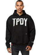 Eine Person mit kurzen Haaren und neutralem Gesichtsausdruck steht vor der Kamera, die Hände an der Kängurutasche, und trägt den trueprodigy Janne Oversized Hoodie mit Print in Schwarz mit fettem "TPDY"-Schriftzug auf der Vorderseite.