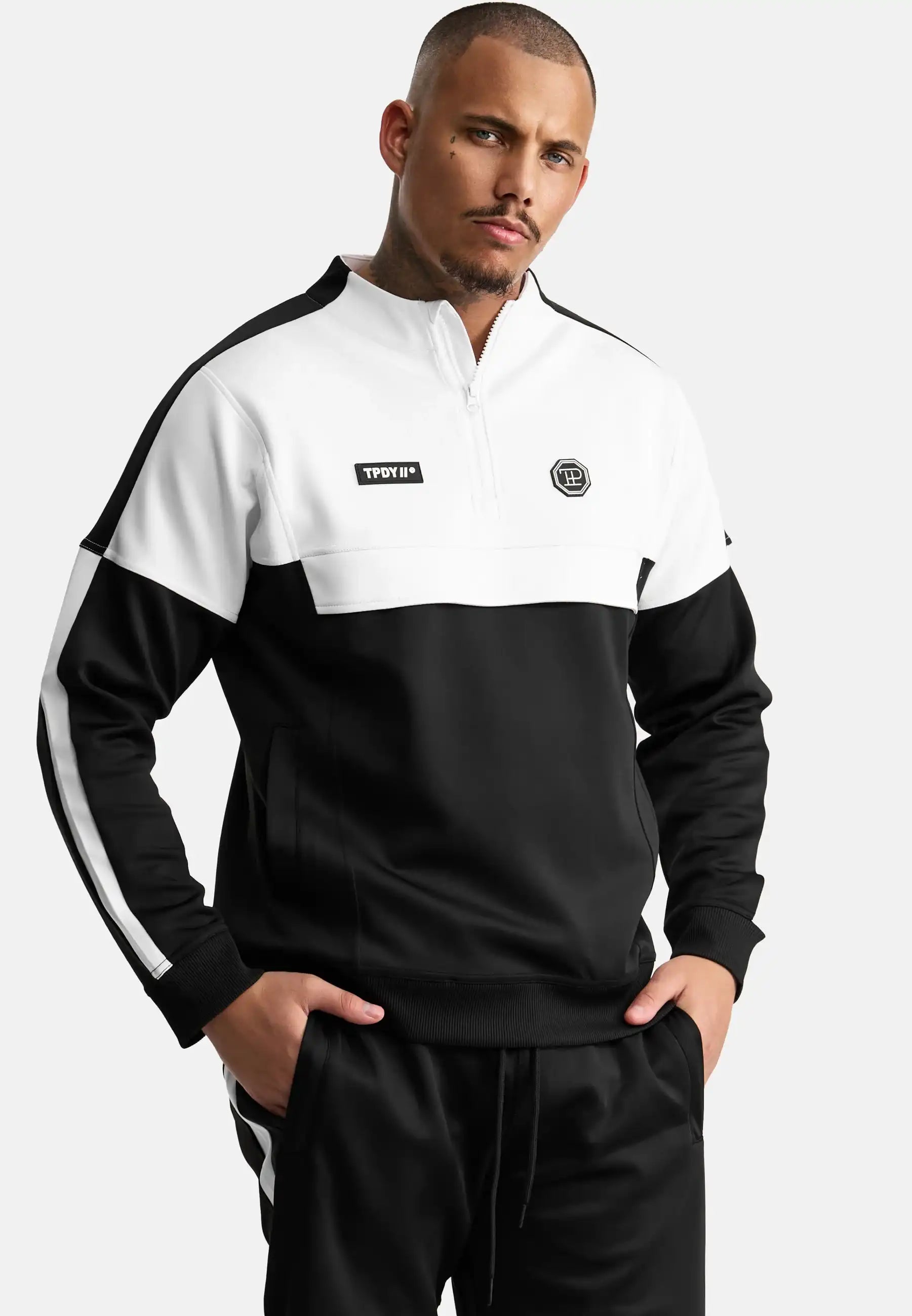 Ein Mann steht mit den Händen in den Taschen und trägt ein trueprodigy Owen Sweatshirt mit Reißverschluss und eine passende schwarze Hose. Das Sweatshirt hat einen Aufnäher mit Text und ein kleines rundes Logo auf der Brust.