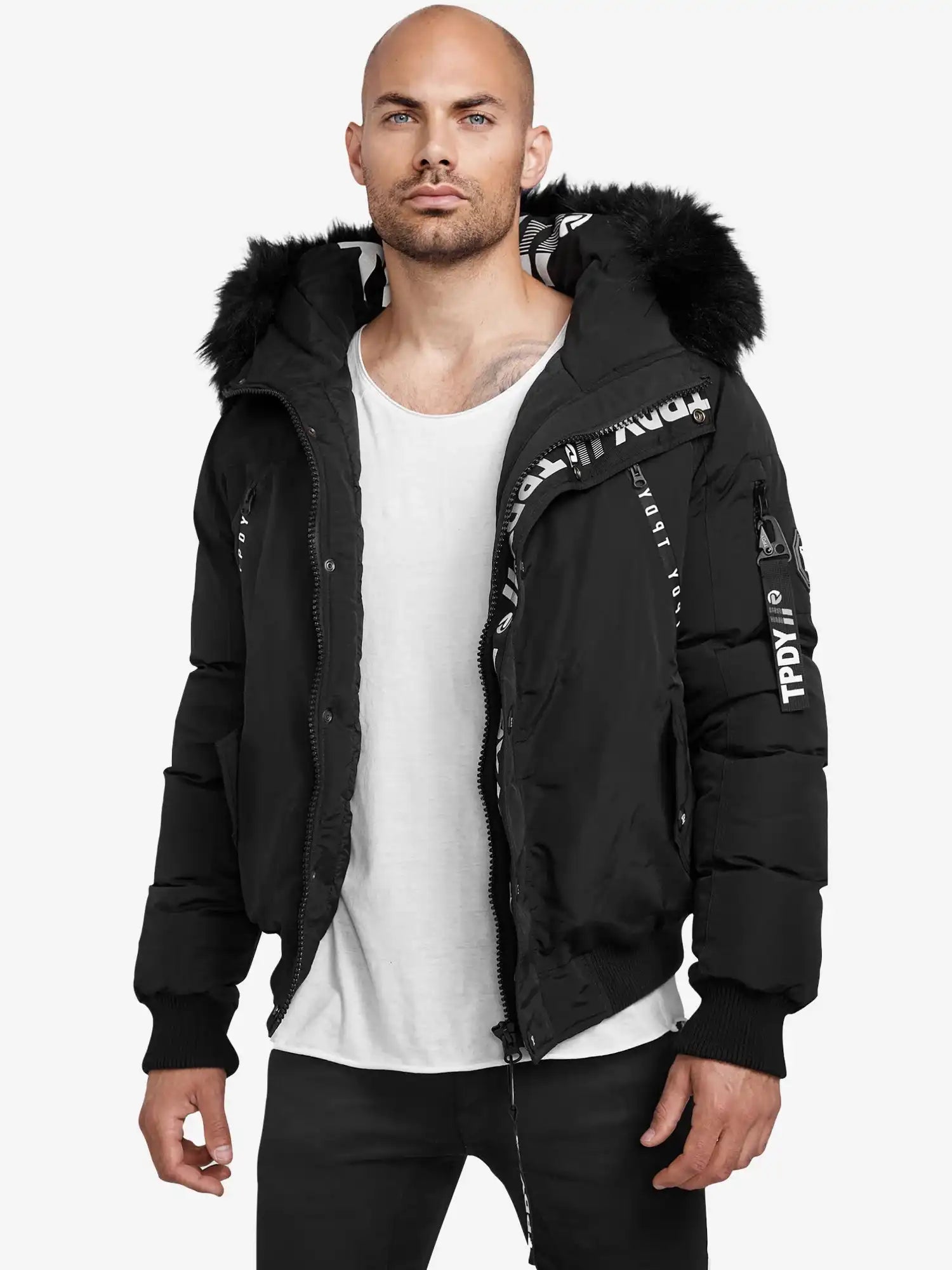 Ein Mann mit rasiertem Kopf und gestutztem Bart trägt die Trueprodigy Noah Winterparka mit Kapuze und Kunstfell, die teilweise geöffnet ist, um weiße Textdetails zu enthüllen, gepaart mit einem weißen T-Shirt und einer schwarzen Hose.