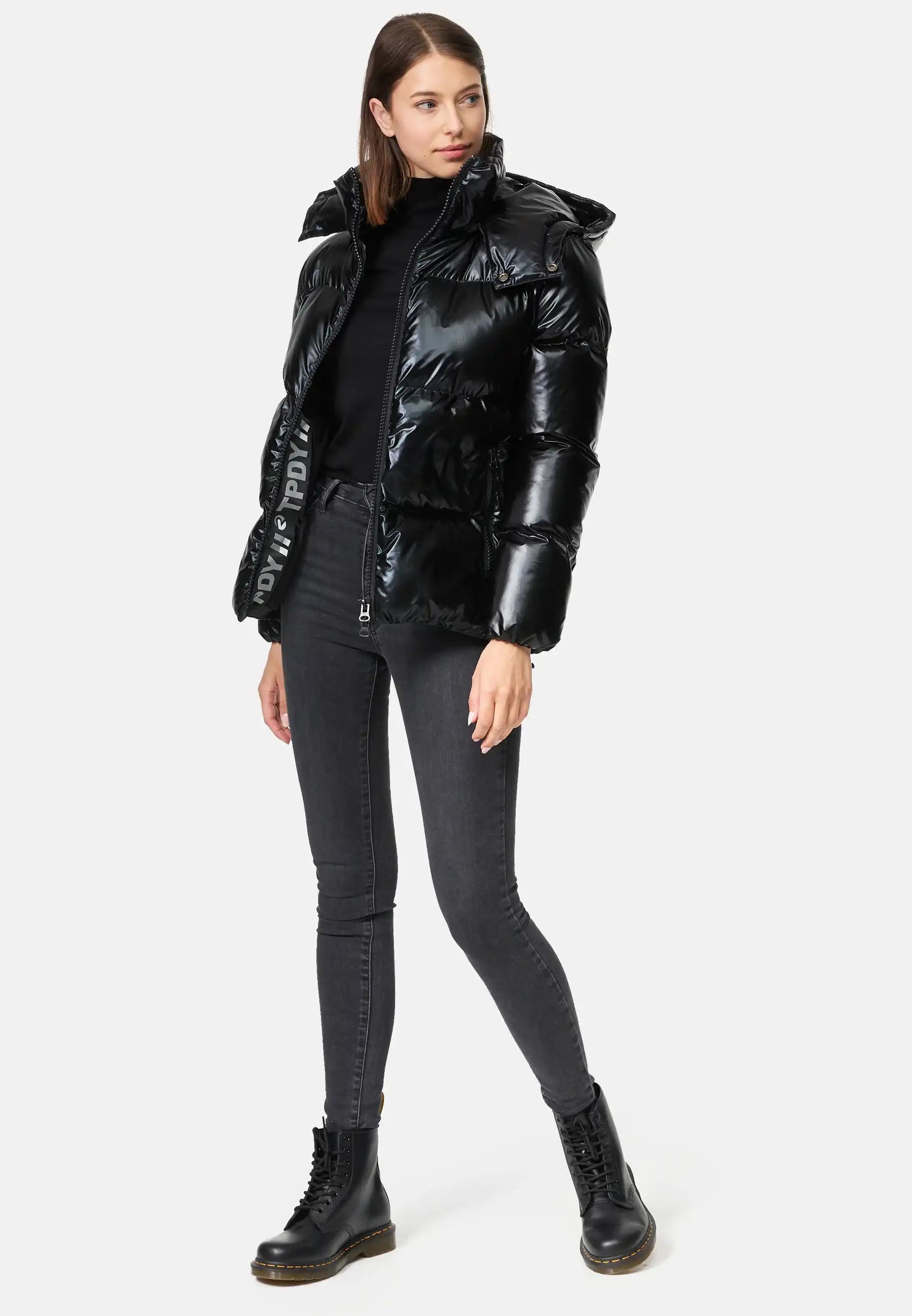 Frau trägt schwarze TRUEPRODIGY constance Pufferjacke mit glänzendem Finish und Kapuze, weißer Schriftzug "TDPDY" am Reißverschluss, kombiniert mit grauen Skinny-Jeans und schwarzen Stiefeln; modischer Winterlook.