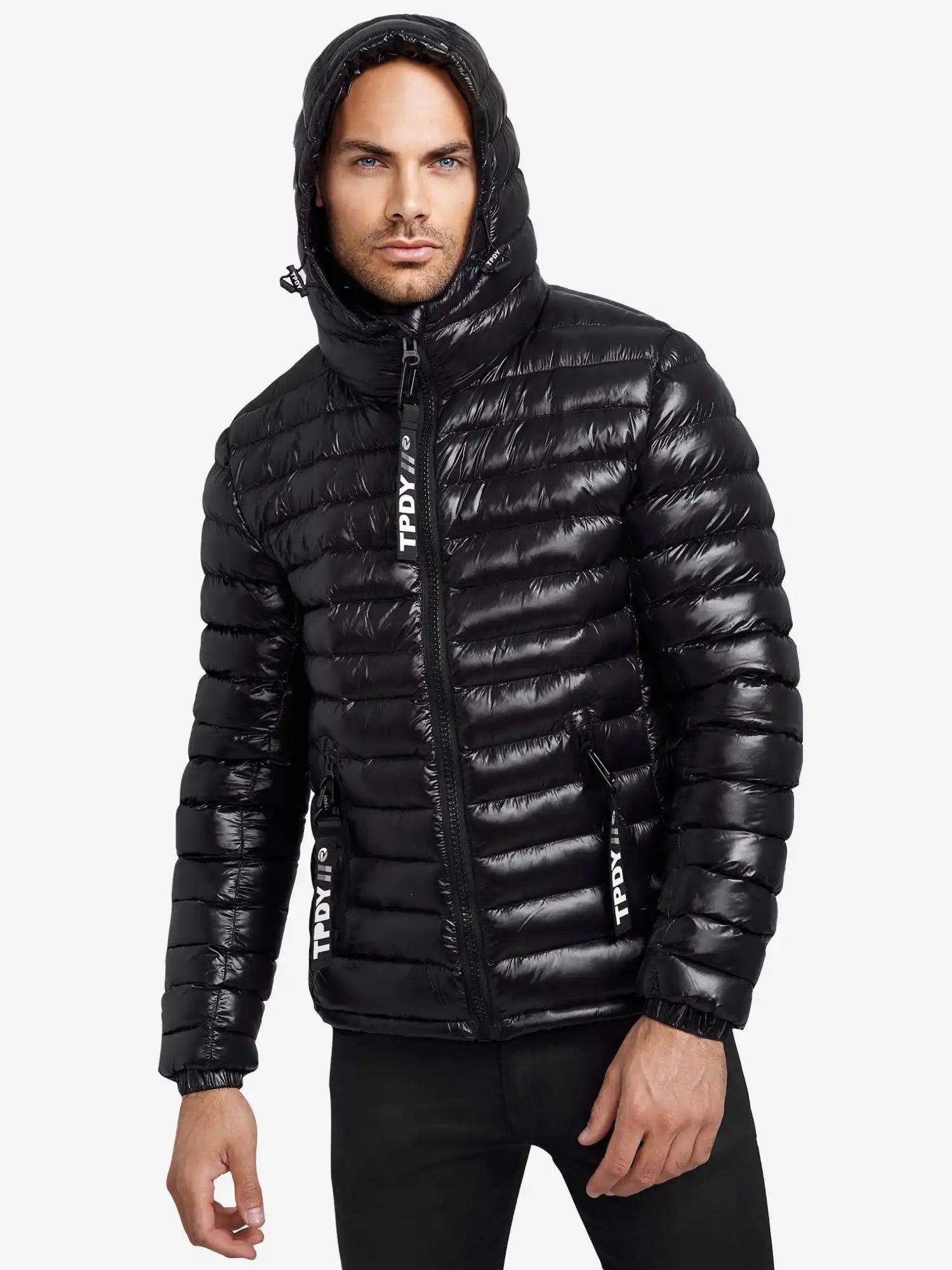 Ein Mann trägt die Adan Steppjacke mit Kapuze von trueprodigy-eine schwarze, wasserabweisende Herrenjacke mit weißen Schriftzügen an den Reißverschlüssen. Er steht vor weißem Hintergrund und blickt seitlich neutral.