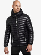 Ein Mann blickt nach vorne in einer glänzend schwarzen trueprodigy Adan Steppjacke mit Kapuze, die mit einer Kapuze, einem Reißverschluss auf der Vorderseite, einer wasserabweisenden Ausrüstung und weißem TPDIVI-Text auf den Reißverschlussleisten vor einem schlichten weißen Hintergrund ausgestattet ist.