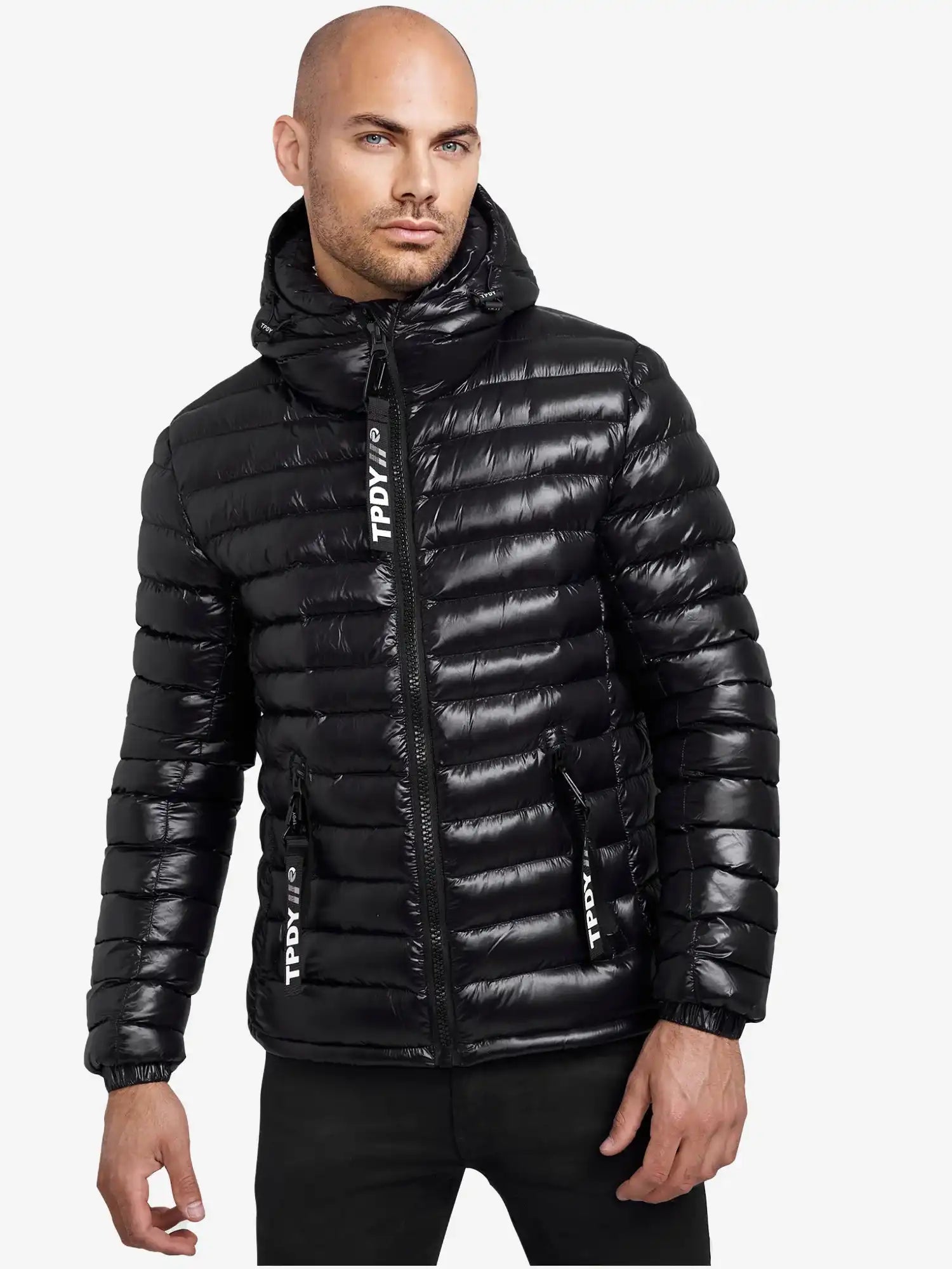 Ein Mann blickt nach vorne in einer glänzend schwarzen trueprodigy Adan Steppjacke mit Kapuze, die mit einer Kapuze, einem Reißverschluss auf der Vorderseite, einer wasserabweisenden Ausrüstung und weißem TPDIVI-Text auf den Reißverschlussleisten vor einem schlichten weißen Hintergrund ausgestattet ist.