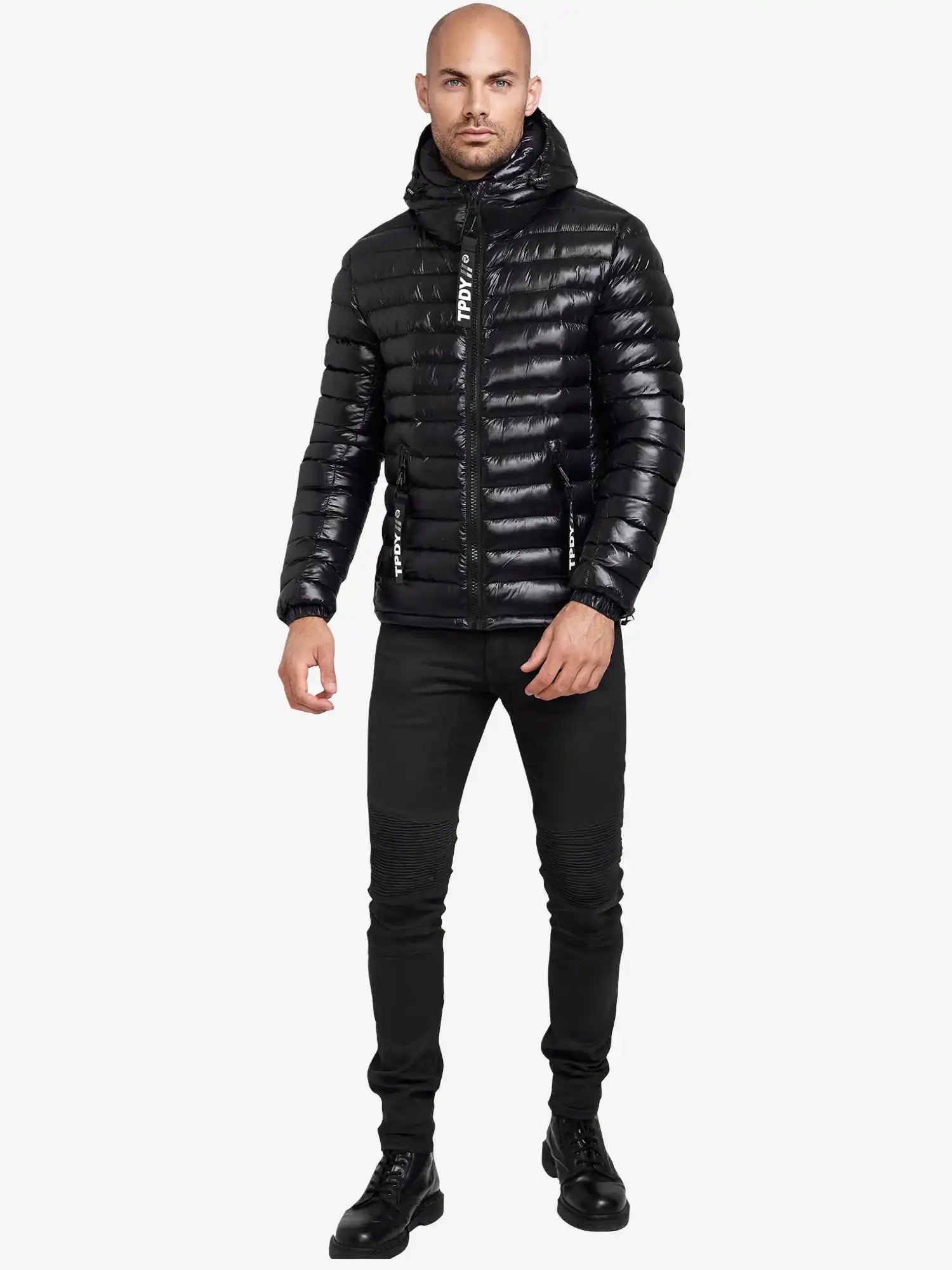 Ein Mann steht mit Blick nach vorne und trägt die glänzende, wasserabweisende schwarze "Adan Steppjacke mit Kapuze" von trueprodigy, gepaart mit einer schmal geschnittenen schwarzen Jeans und schwarzen Schnürstiefeln vor einem schlichten weißen Hintergrund.