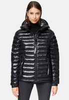 Eine Frau in einer glänzenden schwarzen Adan F Steppjacke mit Kapuze von trueprodigy mit Frontreißverschluss und zwei Seitentaschen steht vor einem weißen Hintergrund und blickt direkt in die Kamera. Die Jacke ist wasserabweisend.