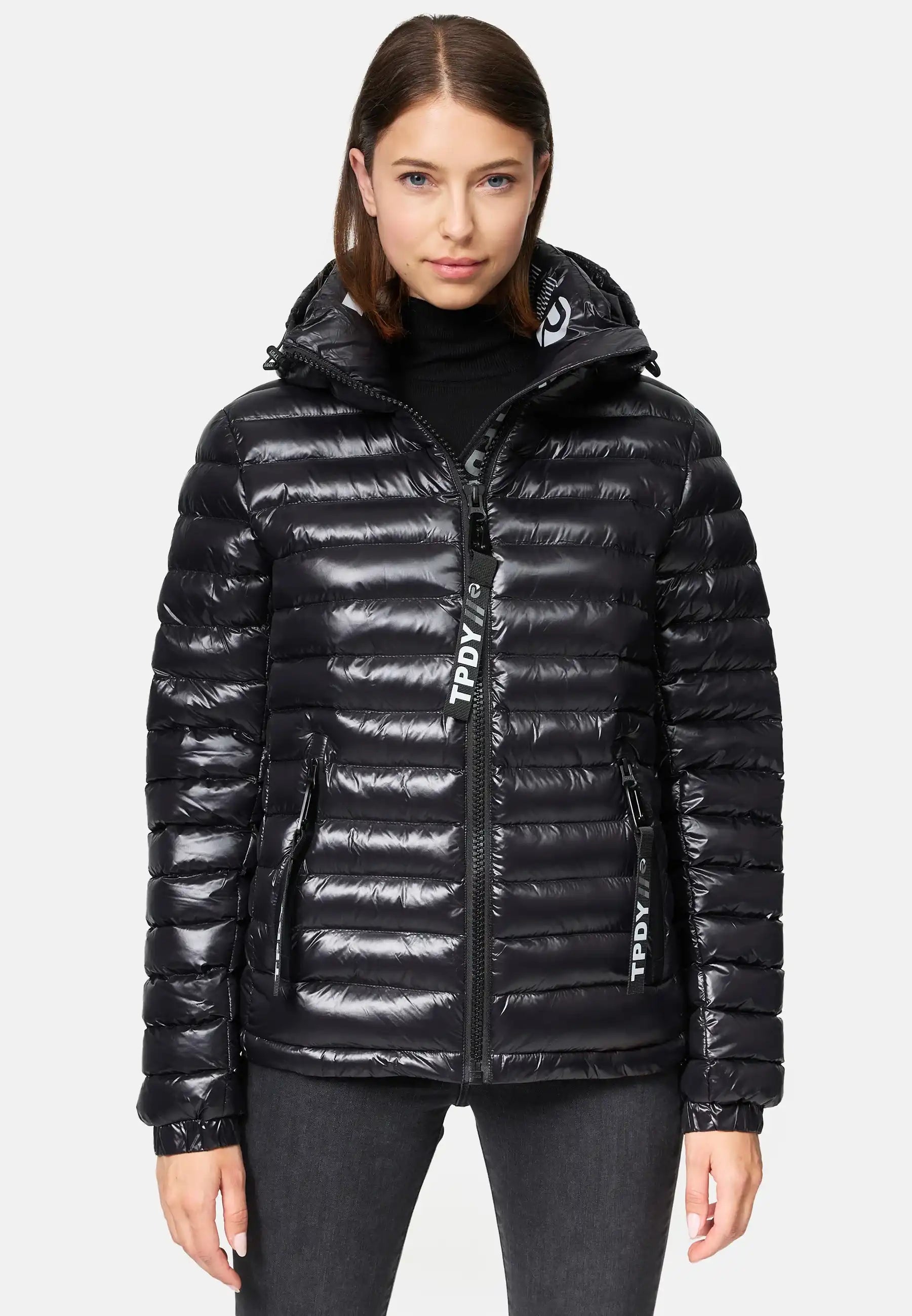 Eine Frau in einer glänzenden schwarzen Adan F Steppjacke mit Kapuze von trueprodigy mit Frontreißverschluss und zwei Seitentaschen steht vor einem weißen Hintergrund und blickt direkt in die Kamera. Die Jacke ist wasserabweisend.