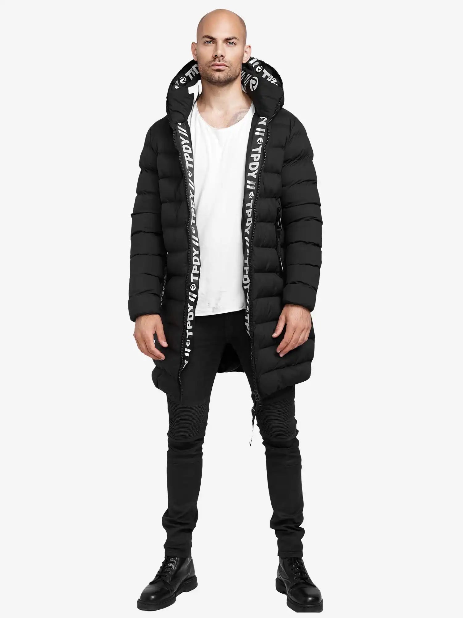 Ein Mann trägt die Chester Lange Pufferjacke mit Logo von trueprodigy in Schwarz mit Kapuze, kombiniert mit weißem T-Shirt, schwarzen Skinny-Jeans und Boots. Die Jacke ist wasserabweisend und zeigt ein Logo auf dem Kapuzenfutter.