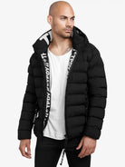 Ein Mann trägt eine schwarze Neo Kurze Pufferjacke mit Logo von trueprodigy mit Kapuze, dazu ein weißes T-Shirt und schwarze Hose, steht vor weißem Hintergrund und blickt nach links.