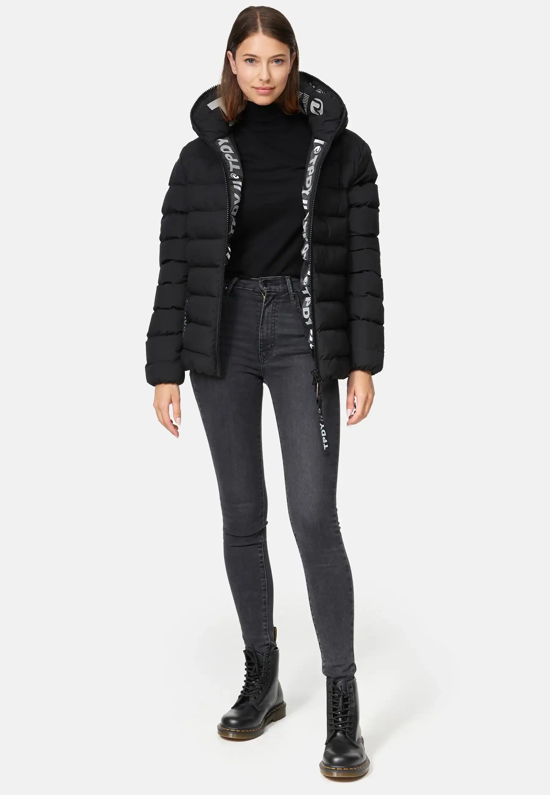 Eine Frau trägt die Neo F Kurze Pufferjacke mit Logo von trueprodigy, kombiniert mit schwarzem Rollkragenpullover, grauen Skinny-Jeans und schwarzen Schnürstiefeln vor hellem Hintergrund.