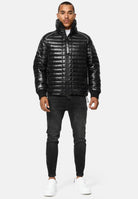 Ein Mann sieht nach vorne und trägt eine glänzende schwarze Corey Steppjacke mit Stehkragen, schwarze Skinny Jeans und weiße Turnschuhe vor einem schlichten hellgrauen Hintergrund.