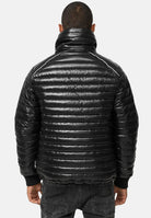 Eine Person ist von hinten zu sehen, die eine glänzende schwarze Trueprodigy Corey Steppjacke mit Stehkragen mit horizontaler Steppung, hohem Kragen und gerippten Bündchen trägt, gepaart mit schwarzen Jeans vor einem schlichten hellen Hintergrund.