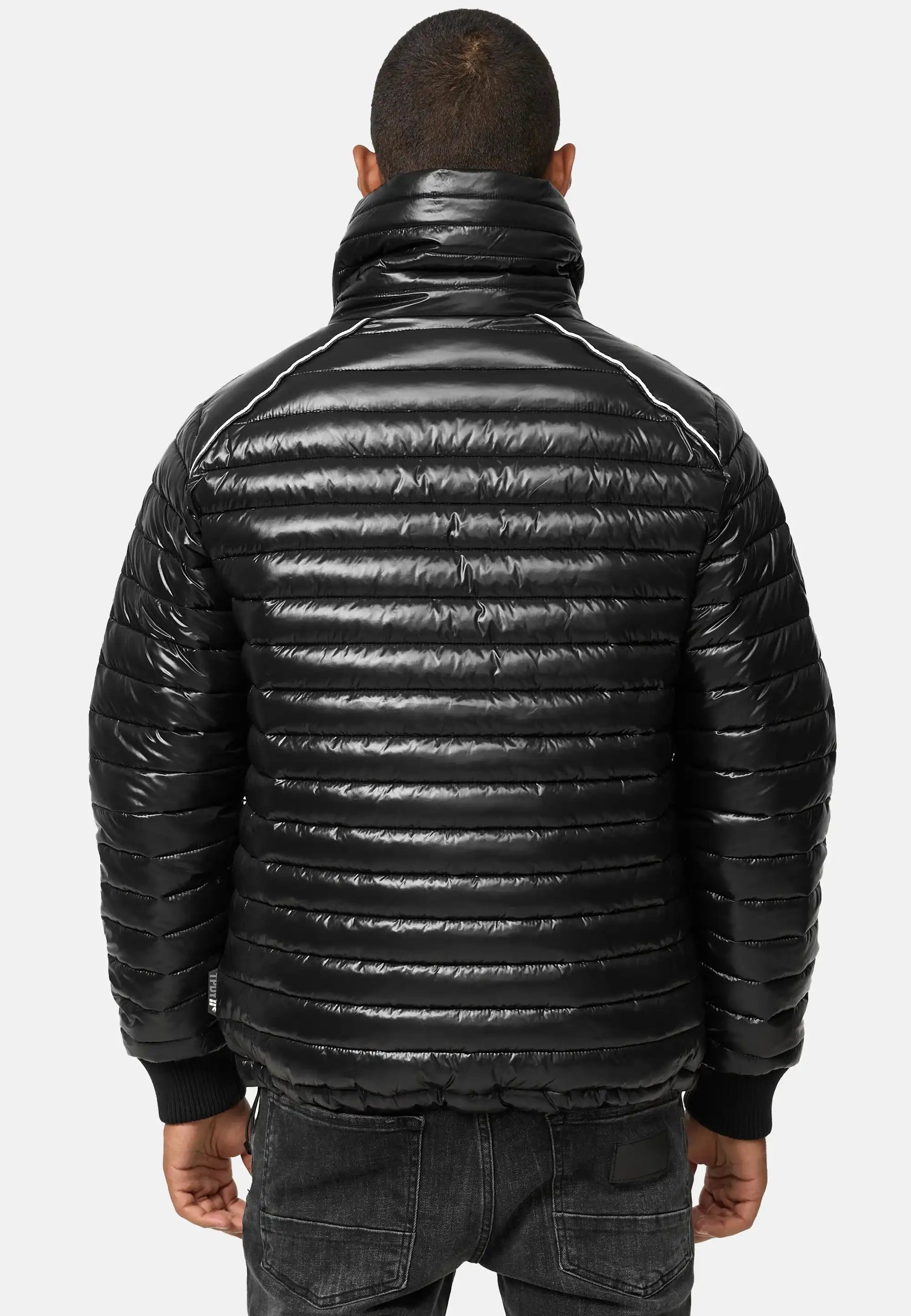 Eine Person ist von hinten zu sehen, die eine glänzende schwarze Trueprodigy Corey Steppjacke mit Stehkragen mit horizontaler Steppung, hohem Kragen und gerippten Bündchen trägt, gepaart mit schwarzen Jeans vor einem schlichten hellen Hintergrund.